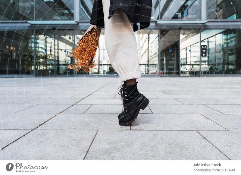 Crop woman with bouquet tiptoeing on street flower pavement style balance urban building female boot pants coat sidewalk glass wall contemporary downtown