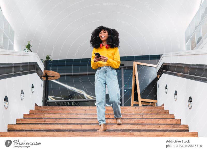 Cheerful stylish ethnic lady walking downstairs and using smartphone in contemporary building woman smile communicate confident trendy positive happy staircase