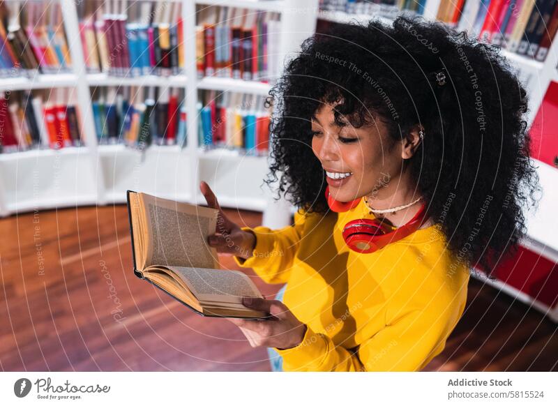 Content ethnic female student reading textbook in library woman smile education happy bookworm bookcase positive trendy young curly hair casual knowledge style