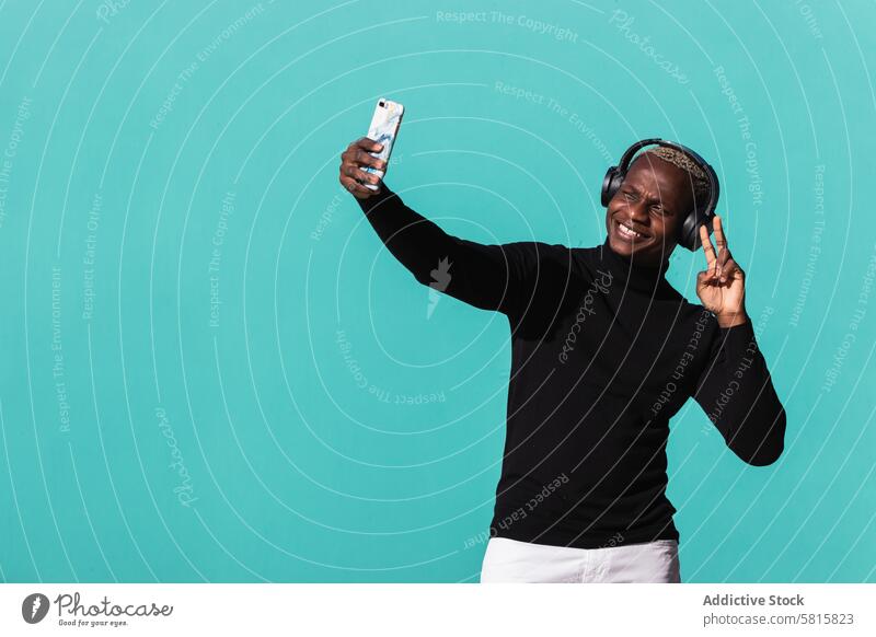 Cheerful young black man listening to music in headphones and taking selfie on street two fingers smile smartphone delight cheerful joy trendy male ethnic