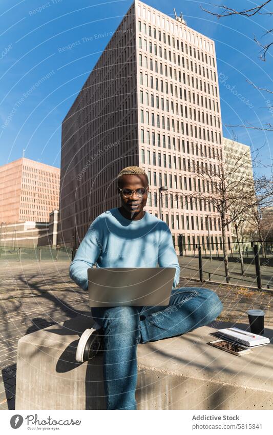 Stylish young black man working remotely on laptop in city park confident self employed typing takeaway coffee bench building male african american ethnic