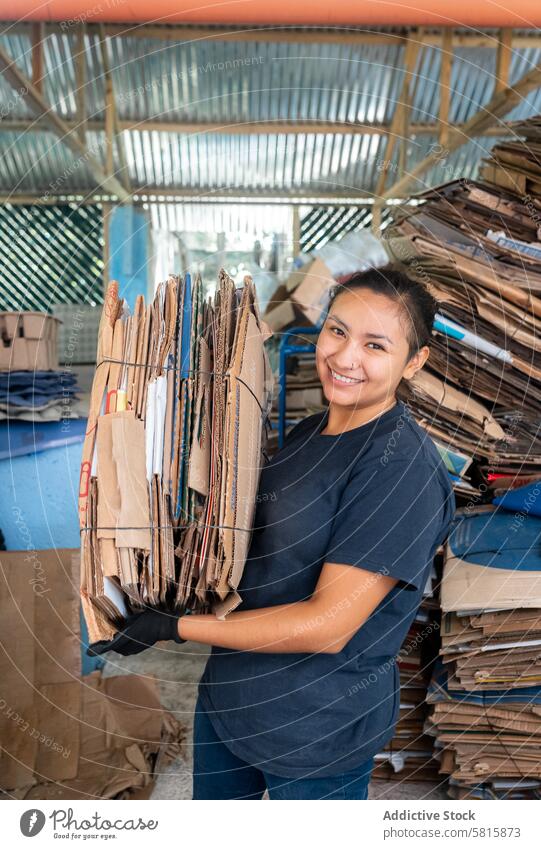 Young Hispanic Woman Smiling While Holding A Heavy Cardboard Pile woman smiling holding heavy cardboard pile happy expression portrait strong focused committed