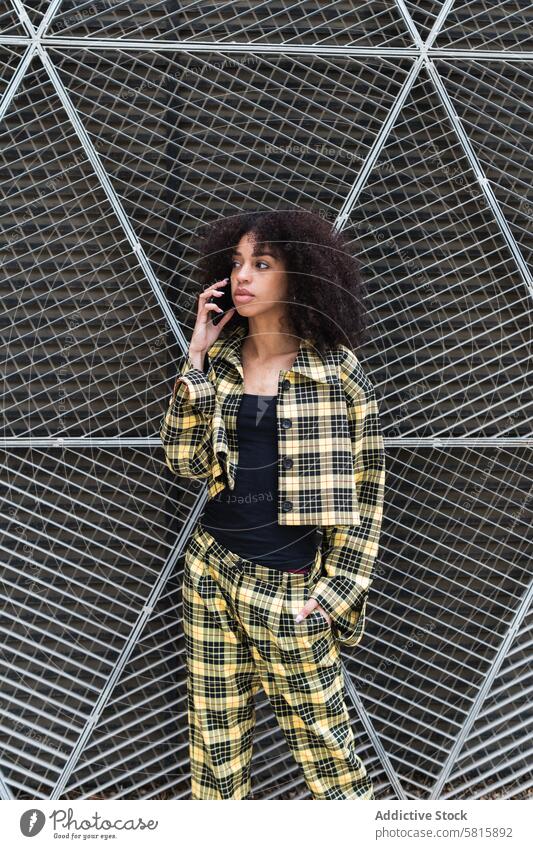 Trendy ethnic woman talking on smartphone against grid fence style fashion hand in pocket individuality cool feminine using gadget device speak cellphone suit