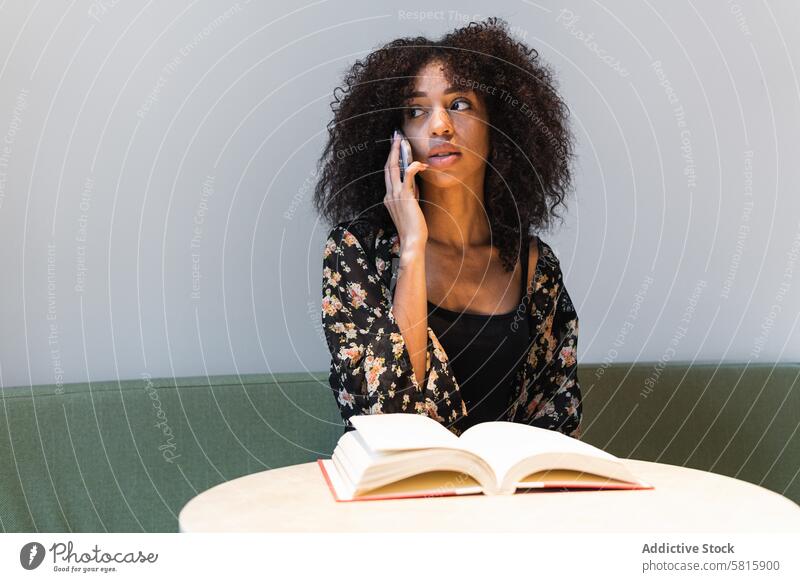 Ethnic woman speaking on smartphone at table with book talk feminine contemplate afro hairstyle sofa using gadget device dreamy cellphone phone call communicate