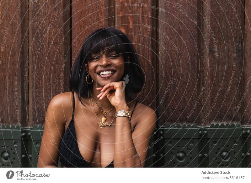 Cheerful black woman in black outfit against wooden wall cheerful happy smile positive glad fun carefree female hand on waist bright pleasure delight joy