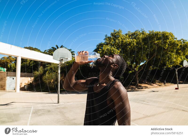 Black fit man drinking water on sunny day on basketball court thirst player sportsman hydrate summer sports ground male ethnic black african american healthy