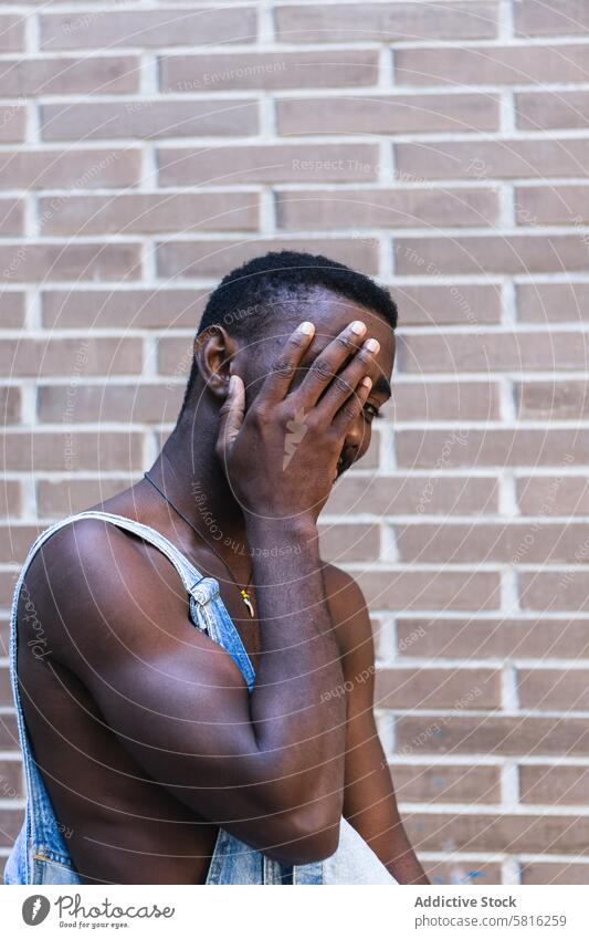 Shy black man on street looking at camera shy cover face handsome overall outfit naked torso smile hide playful male ethnic african american childish young city