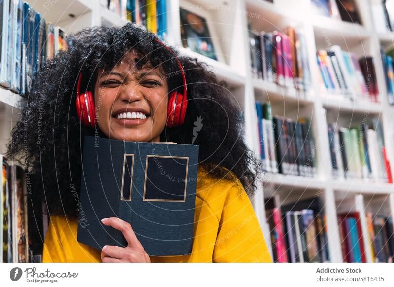 Content ethnic female student with book in library woman listen read music smile education happy bookworm bookcase positive trendy young curly hair casual