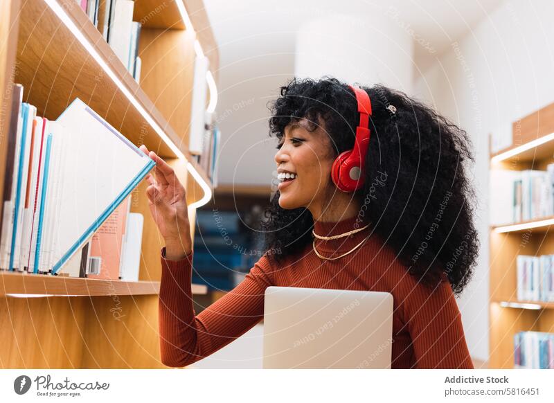 Satisfied woman with headphones taking book in library melomaniac shelf student bookcase positive choose bookshelf smile music listen education literature