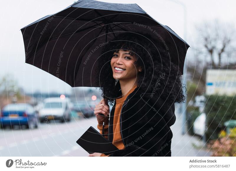 Smiling black woman with umbrella on street rain city roadside urban appearance tablet female portrait african american woman casual young happy positive smile