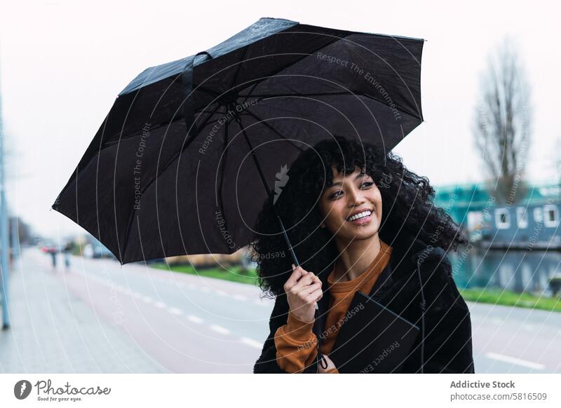 Smiling black woman with umbrella on street rain city roadside urban appearance tablet female portrait african american woman casual young happy positive smile