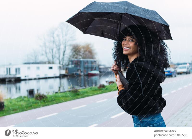 Smiling black woman with umbrella on street rain city roadside urban appearance tablet female portrait african american woman casual young happy positive smile