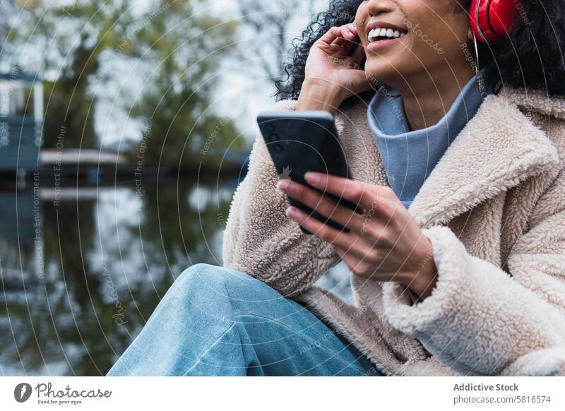 Cheerful black woman listening to music in park headphones smartphone using chill rest relax lake female african american woman city smile positive urban autumn