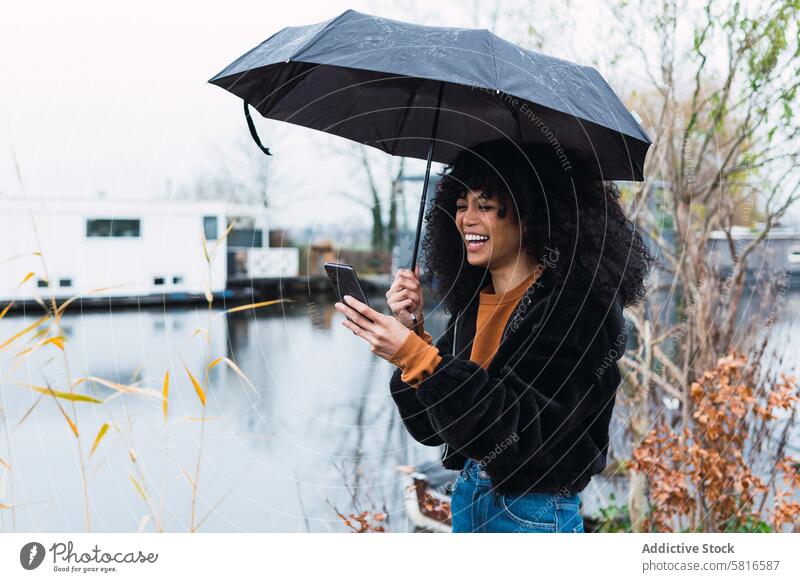 Cheerful black woman with smartphone and umbrella rain city using street urban appearance mobile female african american young happy positive smile cheerful
