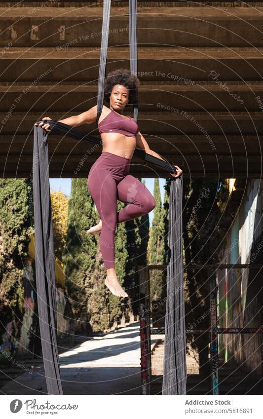 Slim woman doing yoga with aerial silks in city hammock strap balance practice pose acrobatic slender female ethnic african american black hang wellness