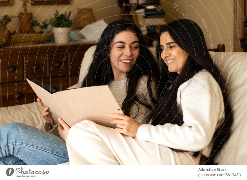 Mature mother and adult daughter smiling while they are watching a photo album home woman together young child happy female people parent house book sitting