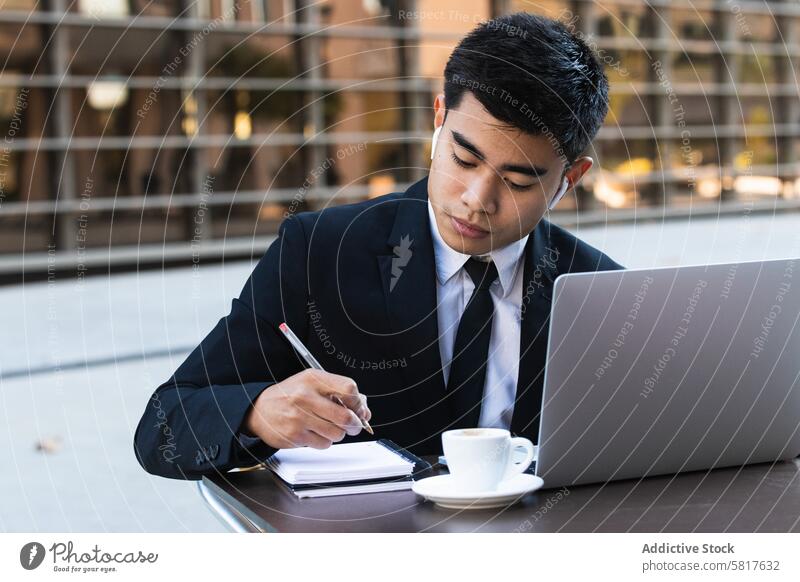 Serious male entrepreneur in suit working in cafe businessman take note notebook remote plan organizer write street ethnic asian notepad earbuds table laptop