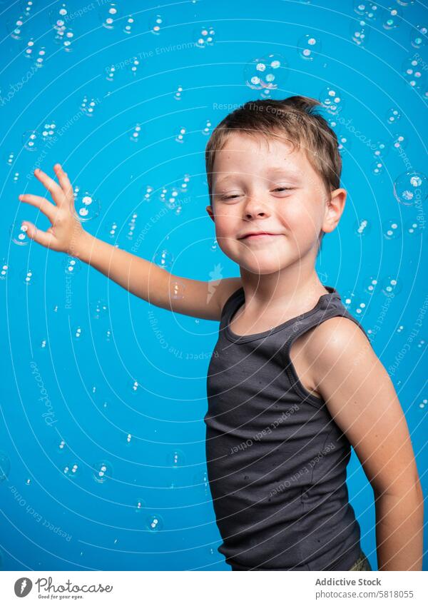 Cheerful boy playing with bubbles on a blue background child studio shot content playful tank top reaching one eye closed looking away joy fun childhood smile