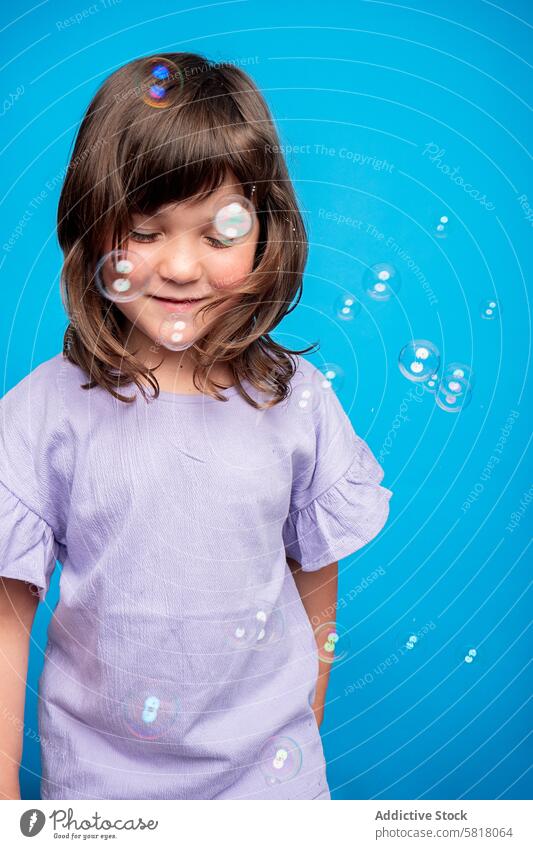 Joyful girl playing with bubbles against blue background child soap fun joy cheerful studio shot purple shirt youth innocence leisure activity indoor playful