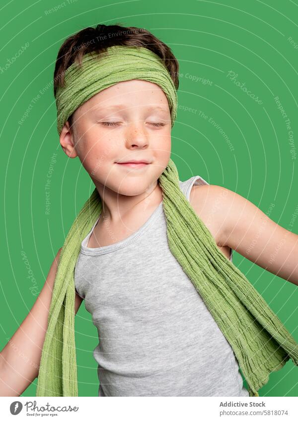 Boy with eyes closed wearing green headscarf, studio shot boy child green background tank top grey content young smiling relaxed fashion style playful innocence