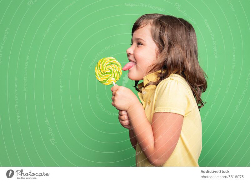 Cheerful girl enjoying a swirl lollipop in studio child cheerful studio shot green background yellow shirt enjoyment candy sweet snack licking treat young