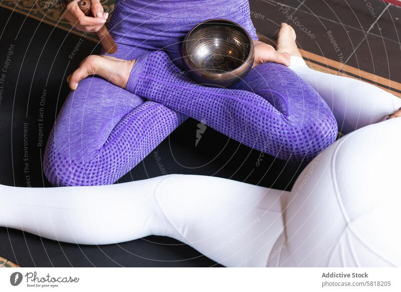 Anonymous woman playing singing bowl during yoga session with female friend women lotus pose padmasana meditate wellness zen tibetan practice calm stress relief