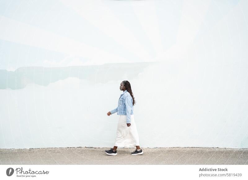 Black Woman in casual wear walking against white wall woman denim jacket white skirt confidence calm peace elegance fashion style outdoor daytime simplicity