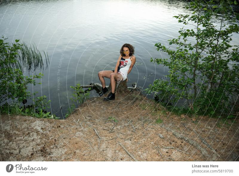 A stunning brunette is enjoying herself in an outdoor forest setting while posing for a model test. This beautiful woman with dark, curly hair, stylish yellow sunglasses, and long legs is looking directly at the camera and exuding a sense of freedom.