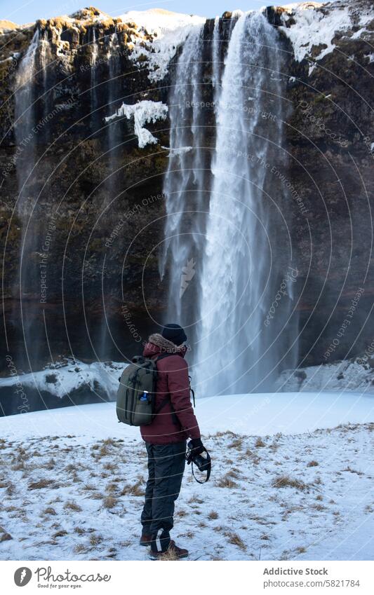 Man with camera facing majestic waterfall in snowy Iceland iceland man photographer backpack nature outdoor winter travel exploration scenic view cold landscape