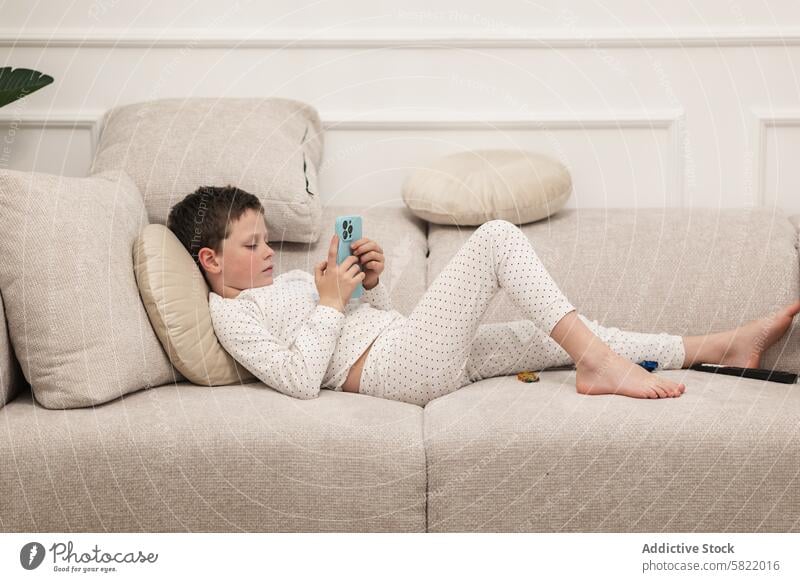 Boy in pajamas using smartphone on couch at home boy child kid polka dot lounge focused indoor lies down technology mobile device screen time comfort casual