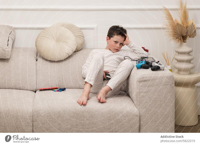 Boy lounging on couch with toys and game controller boy home pajamas bored looking away beige indoor child kid sitting relaxing living room domestic life sad