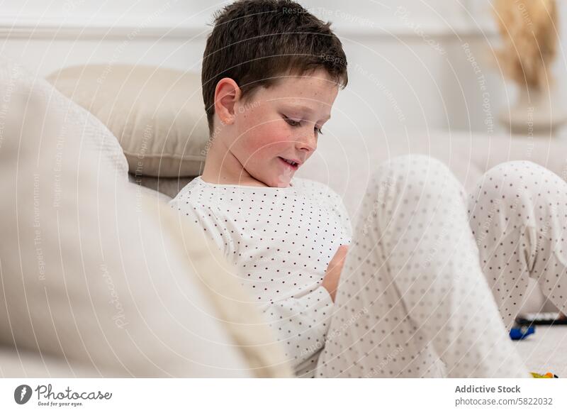 Young boy in pajamas enjoying leisure time at home child sitting couch device handheld gaming looking away comfortable indoor youngster kid entertainment
