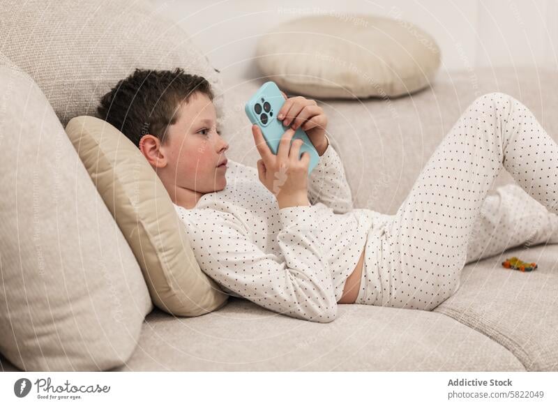 Young boy relaxing with smartphone on couch child kid male home lying absorbed indoor leisure screen technology pajama lifestyle casual comfort living room rest