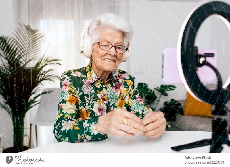 Elderly woman recording creative podcast at home elderly video social media influencer joyful audience headphones shirt floral technology content creator senior