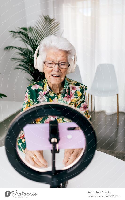 Elderly woman recording a podcast with modern equipment senior elderly technology influencer headphones smartphone ring light smile room bright audio video