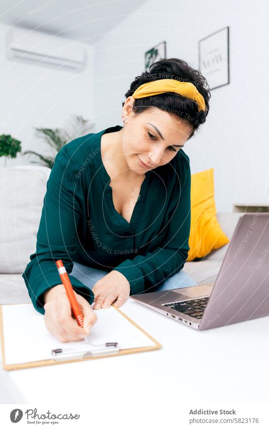 Woman working from home with laptop and clipboard woman writing note seated green blouse bright room decoration focus concentrate indoor modern furniture