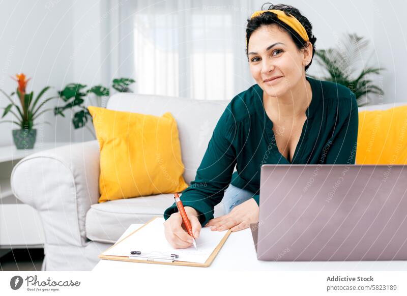Woman writing on clipboard sitting beside laptop at home woman smiling confident green blouse notes colorful pillow camera indoor furniture sofa daylight plant
