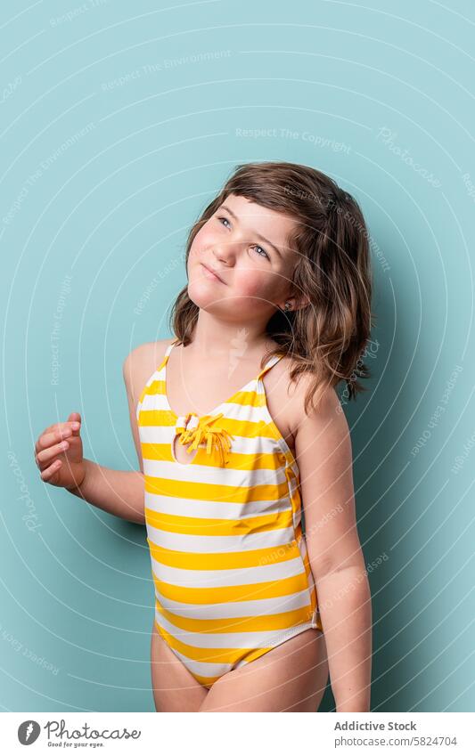 Young girl in striped swimsuit looking up thoughtfully child summer yellow white teal background young pensive standing cute playful innocence vibrant casual