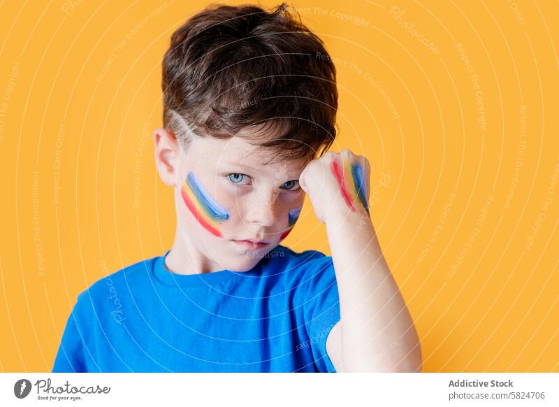 Young boy with rainbow paint on face against yellow background child face paint expression diversity youth color vibrant eye contact pride symbol innocent