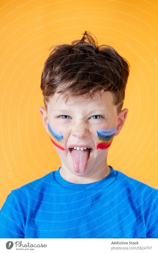 Young boy with colorful face paint sticking out tongue playful orange background blue shirt child rainbow cheek bright joyful cheerful expression young fun