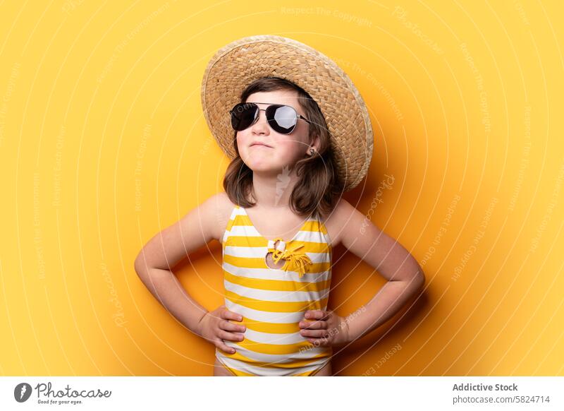 Young girl in a straw hat and sunglasses posing confidently confidence yellow background stripe swimsuit fashion style child pose bright cheerful summer kid