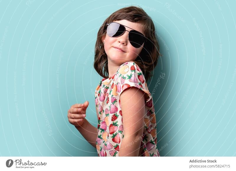 Young girl in sunglasses and fruit-patterned dress poses playfully child smile confident cheerful colorful blue background style fashion kid young cute adorable