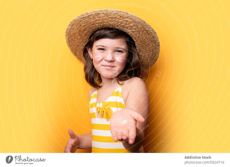 Young girl with sunhat and sunscreen on yellow background straw hat vibrant cheerful swimsuit striped posing summer protection skin care hand offering sunny