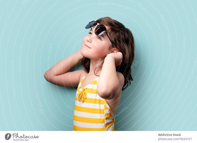 Young girl looking upwards wearing sunglasses young child brunette swimsuit striped yellow white thoughtful gaze teal backdrop summer fashion style cute