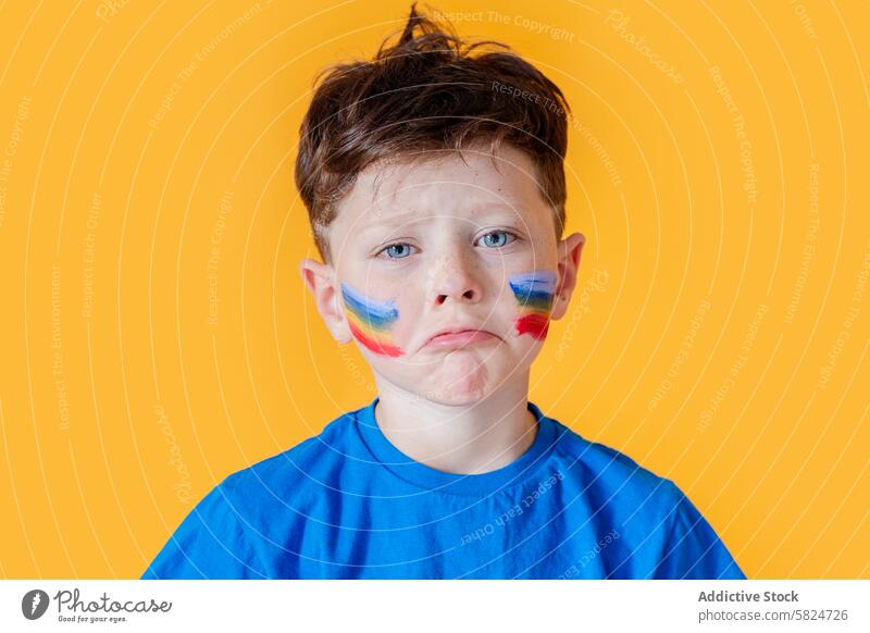 Young boy sad with colorful face paint on yellow background blue curly hair teary vibrant cheek flag expression sadness emotion child kid human face art bright
