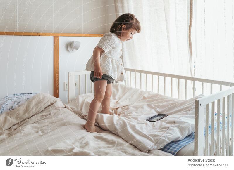 Toddler standing on bed next to crib in a nursery toddler smartphone bedroom child home interior morning pajama daytime life toddlerhood innocence curiosity