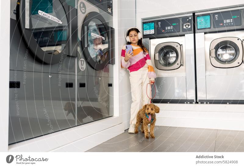 Preteen girl with dog waiting at a laundromat preteen headphones smartphone leash pet washing machine indoors casual clothes laundry multitasking standing