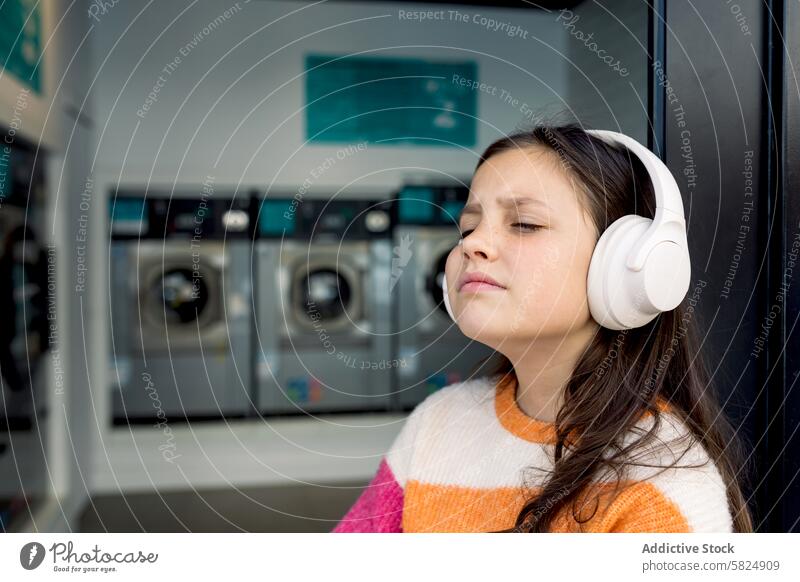 Preteen girl relaxing with headphones in a laundry preteen music eyes closed wearing enjoying backdrop washing machines audio content leisure downtime indoor