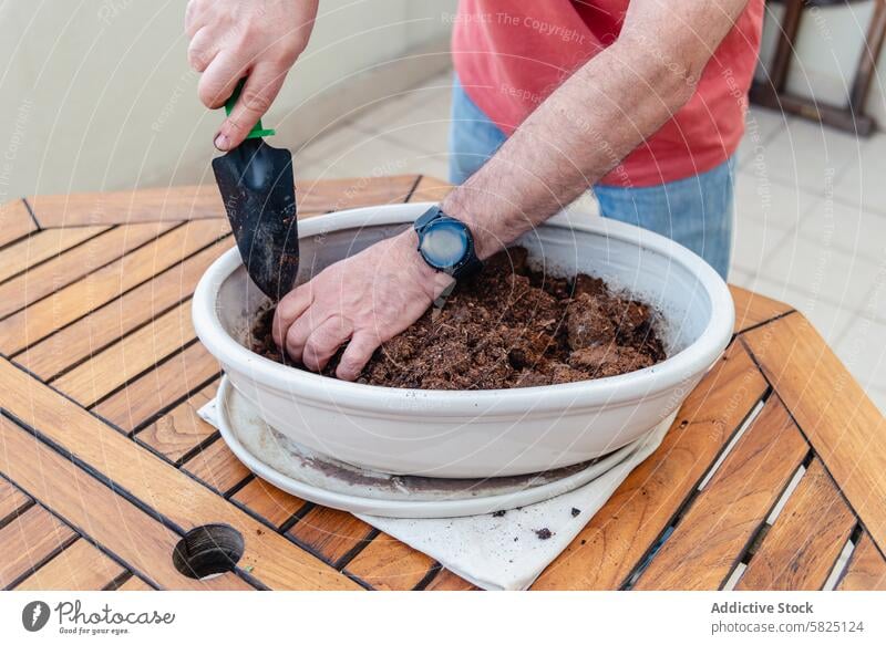 Man planting in a white container on a wooden table man trowel soil outdoor gardening hobby nature wristwatch casual balcony terrace urban green activity hand