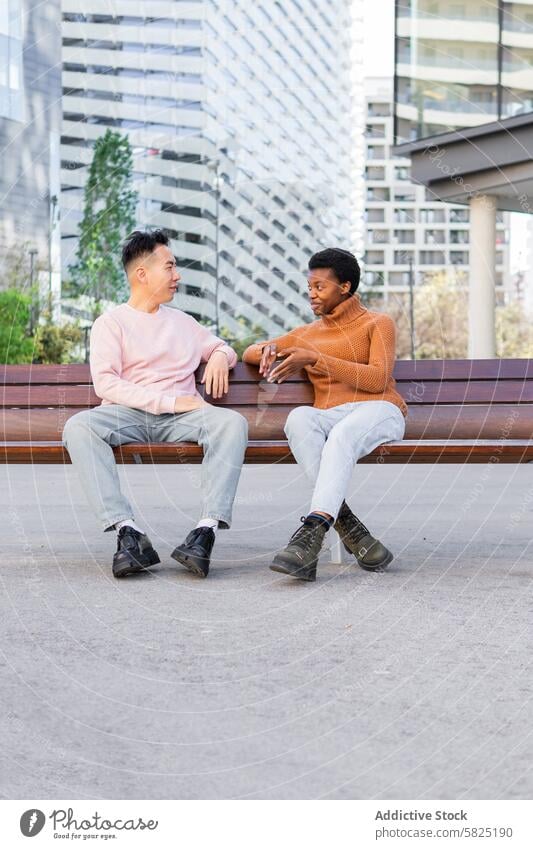 Two young friends chatting on urban bench conversation outdoor asian man african american friendly dialogue sitting multicultural diversity interact city